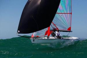 A sailboat with a black sail and two people onboard glides over a green wave on a sunny day, with blue sky in the background.