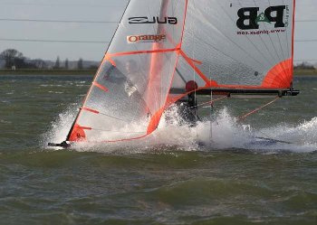A person is sailing on a small boat with an orange and clear sail, cutting swiftly through choppy water and creating a splash. The shoreline and trees are visible in the background.