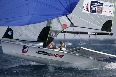 Two sailors compete in a small racing sailboat with 'TEAM GBR' and 'Skandia' logos on the sail. The boat tilts on choppy water during a sailing competition.