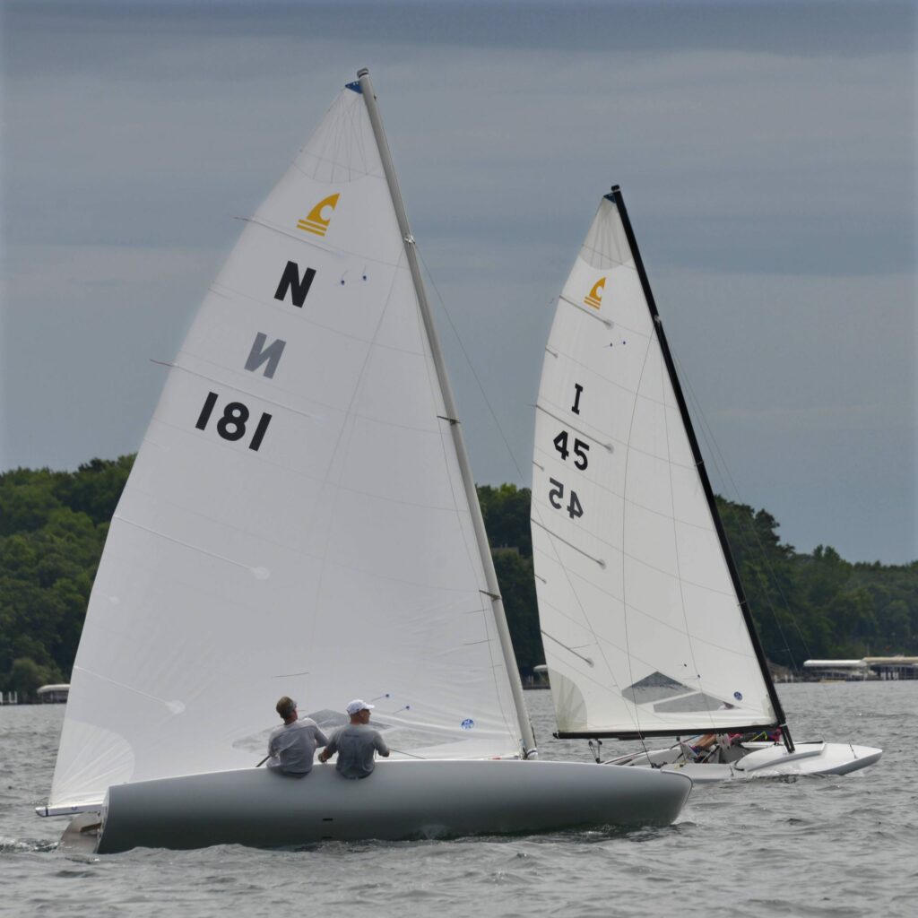 Two people sail a white boat with 'N 181' on the sail, racing alongside another white sailboat marked '45.' Both boats are on a lake with trees and buildings visible in the background under a cloudy sky.