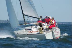 A group of people wearing life jackets sail a white sailboat on choppy blue water under a clear sky. The boat is tilting as it moves quickly, and the shoreline is visible in the distance.