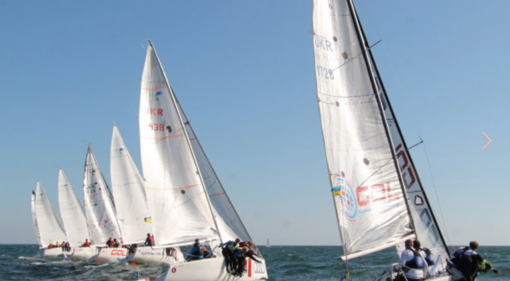 Several sailboats with crews race closely together on open water under a clear blue sky, their white sails catching the wind.