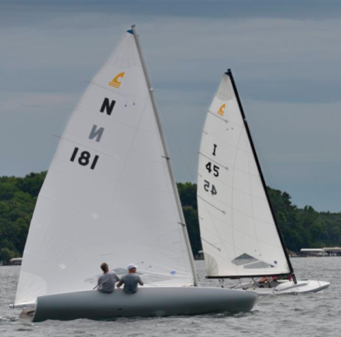 Two sailboats with white sails, each carrying two people, race on a lake under a cloudy sky. Trees and a few lakeside buildings are visible in the background.
