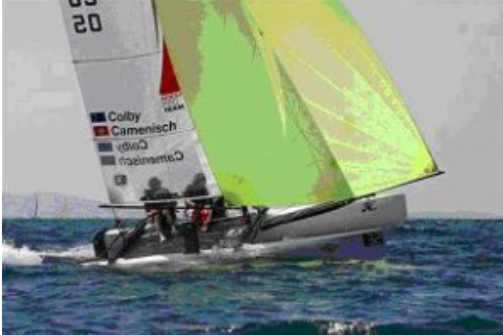 A small sailboat with a neon yellow sail glides across the ocean, carrying several crew members dressed in dark gear. The boat features visible sponsor logos and names on the sail.