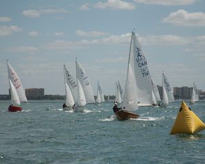 Several sailboats with white sails race on a body of water near a yellow buoy, with city buildings visible in the background under a partly cloudy sky.