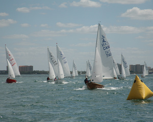 Several sailboats with white sails race on a body of water near a yellow buoy, with city buildings visible in the background under a partly cloudy sky.