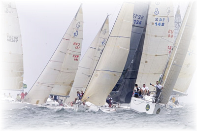 Several sailboats with crews compete in a close race on the water, their sails tilted in the wind under an overcast sky.
