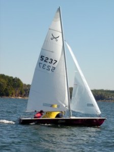 A small sailboat with two people onboard sails on a lake. The boat has white sails marked with the numbers '5237.' Trees and shoreline are visible in the background under a clear sky.