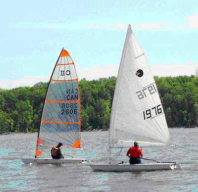 Two people sail separate small boats on a lake, one with a blue and orange sail and the other with a white sail, with a forested shoreline and cloudy sky in the background.
