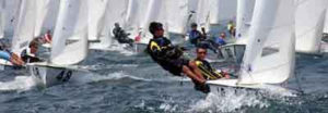 Several teams of sailors race small sailboats on choppy water; two sailors in the foreground lean out of their boat to balance it. Numerous white sails fill the background under a sunny sky.