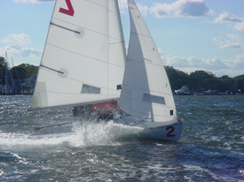 A small sailboat with the number 2 on its bow moves quickly through choppy water, creating a splash. The boat has two sails and there are trees and other boats in the background under a partly cloudy sky.