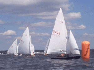 Several sailboats with white sails are racing on a body of water, rounding an orange buoy on a partly cloudy day. The water appears choppy and the boats are closely grouped together.