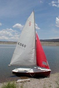 A small sailboat with a white and red sail is docked on the shore of a calm lake under a partly cloudy sky. The boat is resting on shallow water near a rocky shoreline.