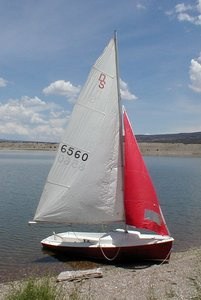 A small sailboat with a white and red sail is docked on the shore of a calm lake under a partly cloudy sky. The boat is resting on shallow water near a rocky shoreline.