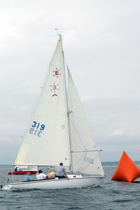 A small sailboat with three people on board sails near an orange buoy on a cloudy day, with a red boat visible in the background.