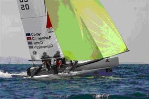 Two people sail a small racing catamaran with a white hull and neon yellow sail over blue water. The sail displays names 'Colby' and 'Camenisch' along with racing numbers.