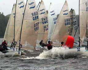 Several sailboats with numbers and country codes compete closely on choppy water, with sailors maneuvering near a red buoy. Trees are visible in the background under a cloudy sky.