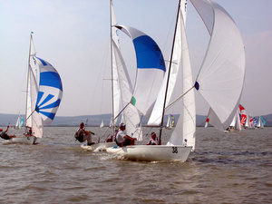 Several small sailboats with colorful sails race on a lake, with people actively steering. Other sailboats are visible in the background under a partly cloudy sky.
