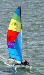 A person sails a small catamaran with a colorful, rainbow-striped sail on choppy water. The sail displays the numbers 1077 and the letters MAR CAN.