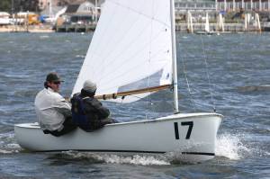 Two people sail a small white sailboat with the number 17 on the bow across choppy water, with buildings and other boats visible in the background.