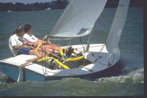 Two people are sailing a small sailboat on a body of water. They are leaning back to balance the boat, which is tilted due to the wind in its sails. The water shows slight waves and the sky is clear.