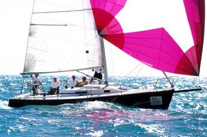A sailboat with four people aboard glides over blue ocean waves. The boat has white sails and a large, colorful pink and white spinnaker. The sun is shining brightly, creating sparkling reflections on the water.