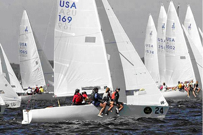 Several sailboats with white sails, marked with 'USA' and numbers, race on choppy water. Crew members in life jackets are actively working on the boats under a cloudy sky.