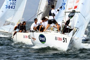 A group of sailors competes in a sailboat race, navigating choppy water. The crew members wear white shirts and shorts, and the boats display various sponsor logos and numbers on their sails and hulls.