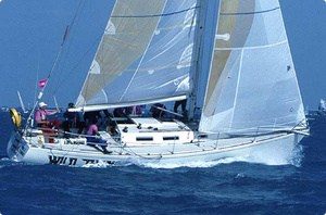 A white sailboat with several people on board sails quickly over blue ocean water, its sails full of wind against a clear sky.