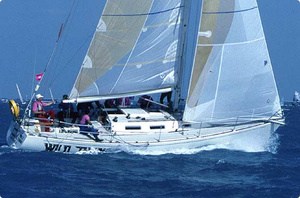 A white sailboat with several people on board sails quickly over blue ocean water, its sails full of wind against a clear sky.