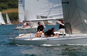 Three people are sailing a small white sailboat in a race, all wearing black shirts. Other sailboats are visible in the water in the background. The weather appears clear and sunny.