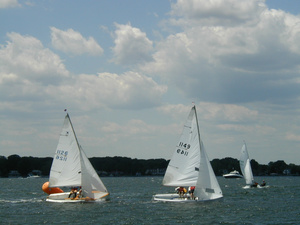 Three sailboats with white sails are racing on a lake under a partly cloudy sky, with a tree-lined shore in the background and a bright orange buoy marking the course.