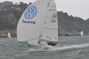 A white sailboat with a large Volkswagen Touareg logo sails on choppy water near a coastline with trees and buildings in the background. Several people are visible on the boat.