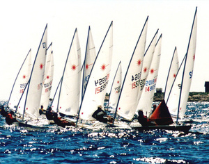 Several small sailboats with white sails race closely together on choppy blue water, with sailors leaning out to balance the boats under a bright sky.