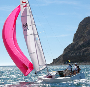A small white sailboat with a bright pink sail glides on the water near a rocky coastline. Three people are on board, enjoying sunny weather. The sea is calm, and the sky is clear.