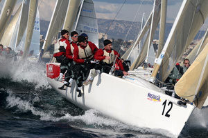 A team of sailors in red and white gear lean over the side of a racing sailboat numbered 12, cutting through choppy water with other sailboats in the background.
