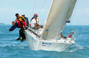 Five people sail a white racing sailboat on blue water, with two crew members leaning out over the side for balance. The boat tilts in the wind, and the crew wears casual sailing gear and hats.