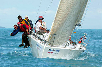Five people sail a white racing sailboat on blue water, with two crew members leaning out over the side for balance. The boat tilts in the wind, and the crew wears casual sailing gear and hats.