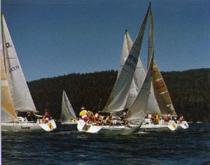 Several sailboats with people wearing colorful life jackets are racing on a body of water, with forested hills and a clear blue sky in the background.