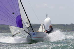 A person sails a small boat with purple and white sails through choppy water, creating splashes. Other sailboats and a green coastline are visible in the background under a bright sky.