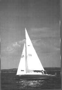A sailboat with two large white sails glides across calm water under a partly cloudy sky, with distant land visible on the horizon.