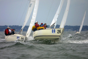 Two sailboats with crews dressed in colorful jackets race side by side on choppy water under a cloudy sky. The boat in front displays the number 26, while the boat behind shows the number 09.