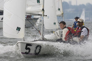 Two people wearing life jackets sail a small white dinghy marked with the number 50 on choppy water, while other boats and sailors are visible in the background.