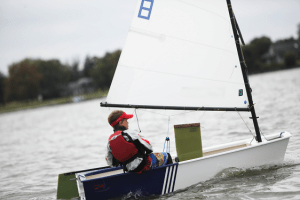 A person wearing a red life jacket and visor sails a small white dinghy on a lake, with trees and houses blurred in the background under a cloudy sky.