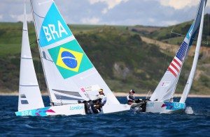 Two sailboats with sailors compete on the water. One boat displays the Brazilian flag and "BRA," while the other shows the U.S. flag and "USA." A coastline and cloudy sky are visible in the background.