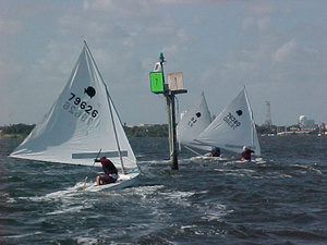 Three small sailboats with white sails navigate choppy waters near a green channel marker with the number 7, under a partly cloudy sky. The sailors are leaning to balance their boats.