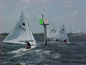 Three small sailboats with white sails navigate choppy waters near a green channel marker with the number 7, under a partly cloudy sky. The sailors are leaning to balance their boats.