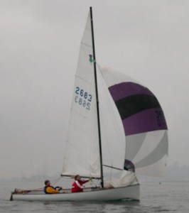 Two people sail a small white sailboat with a tall mast and a purple, black, and white spinnaker on a calm, overcast day. The water is smooth and the background is hazy.