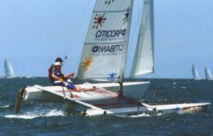 A person sails a small catamaran with colorful sun graphics and sponsor logos on the sail across choppy blue water, with other sailboats visible in the background.