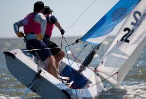 Two people wearing life jackets are sailing a small boat with a blue and white sail on the water, leaning out to balance as the boat tilts in the wind.